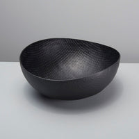 A black aluminum bowl with a textured crosshatch pattern, placed on a white surface against a plain background.